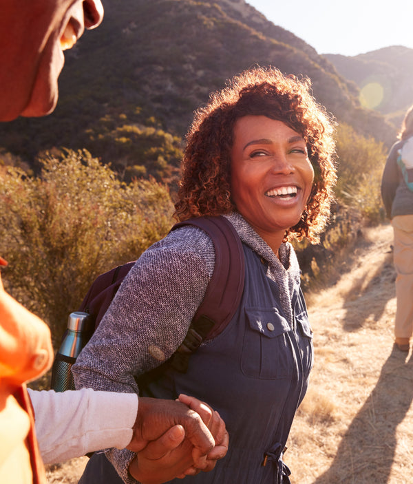 Smiling woman with a backpack hiking a sunlit trail, holding a companion's hand